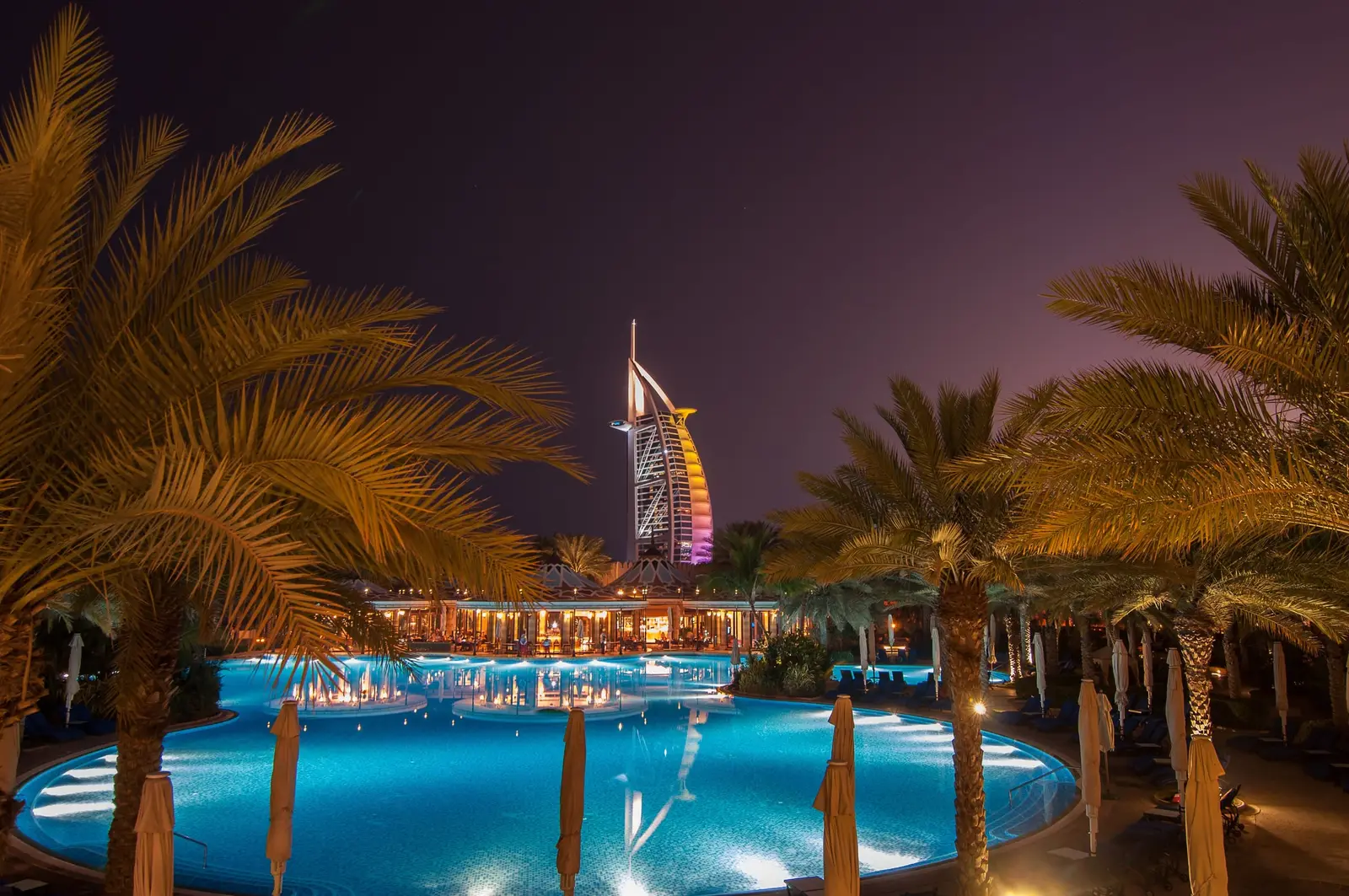 Burj Al Arab lit up behind palm trees and a resort pool at night, Dubai